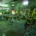 緑の照明に包まれたレトロなカフェで金属のロボットたちがくつろいでいる。